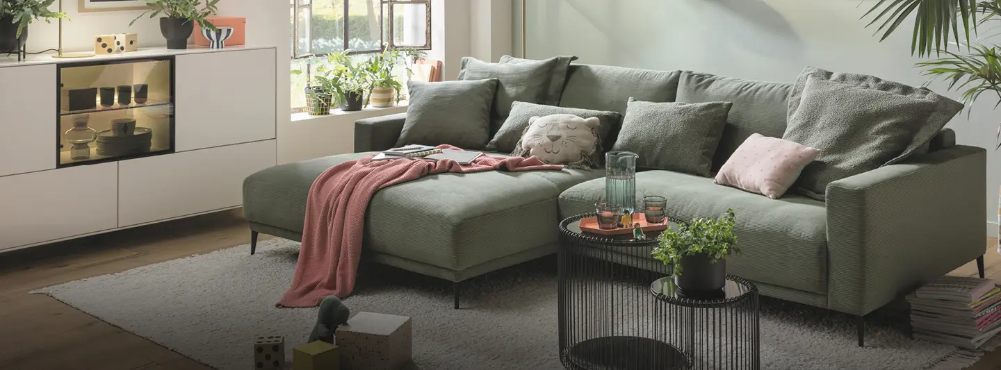 Wohnzimmer mit grünem Ecksofa, Kissen und Decke, davor runder Couchtisch und Teppich, links Sideboard mit Pflanzen